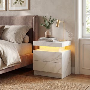Peut inclure: Une table de chevet moderne en imitation marbre blanc, avec deux tiroirs et un éclairage intégré. Une lampe dorée et un petit vase avec de la verdure sont posés dessus. La table de chevet est à côté d'un lit avec une tête de lit et une literie mauves.