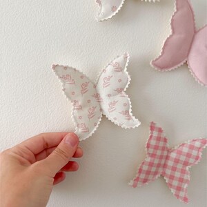 May include: A collection of fabric butterfly decorations in various shades of pink and white. One butterfly is held by a hand. The butterflies have different patterns, including floral prints and gingham. The edges are scalloped.