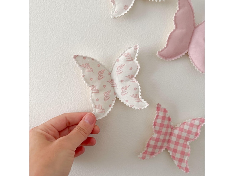 May include: A collection of fabric butterfly decorations in various shades of pink and white. One butterfly is held by a hand. The butterflies have different patterns, including floral prints and gingham. The edges are scalloped.