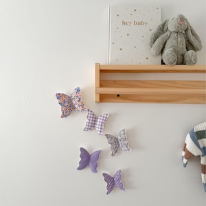 May include: A collection of fabric butterflies in various patterns and colors, including floral, gingham, and solid purple, are arranged on a white wall. A wooden shelf holds a book with the text "hey baby" and a gray stuffed bunny. A striped sweater hangs nearby.