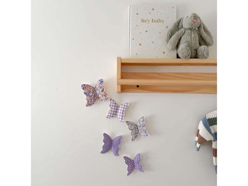 May include: A collection of fabric butterflies in various patterns and colors, including floral, gingham, and solid purple, are arranged on a white wall. A wooden shelf holds a book with the text "hey baby" and a gray stuffed bunny. A striped sweater hangs nearby.