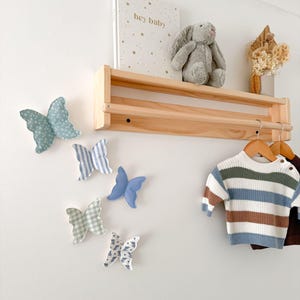 May include: A nursery wall decor scene featuring a wooden shelf with a plush bunny, a book with the text "hey baby", and dried flowers. Decorative fabric butterflies in various patterns and colors are affixed to the wall. A striped sweater hangs on a wooden hanger.
