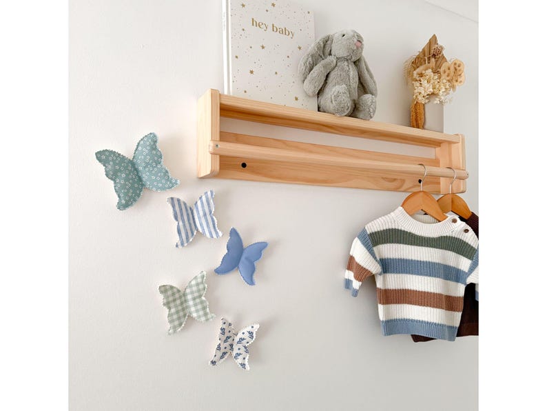 May include: A nursery wall decor scene featuring a wooden shelf with a plush bunny, a book with the text "hey baby", and dried flowers. Decorative fabric butterflies in various patterns and colors are affixed to the wall. A striped sweater hangs on a wooden hanger.