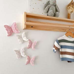 May include: A collection of fabric butterfly wall decorations in various shades of pink and white, with different patterns. A wooden shelf holds a stuffed bunny, a book, and dried flowers. A striped sweater hangs on a wooden hanger.