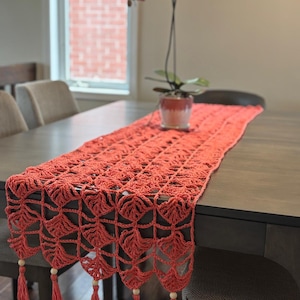 May include: Coral-colored, hand-crocheted table runner with a leaf-like pattern. The runner has a scalloped edge with tassels and wooden beads. A potted orchid sits on the table. This decorative table runner is perfect for adding a touch of handmade charm to any dining space.