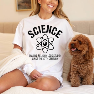 Peut inclure: T-shirt blanc avec le mot "SCIENCE" au-dessus d'un symbole d'atome et le texte "MAKING RELIGION LOOK STUPID SINCE THE 17TH CENTURY" en noir. Un petit chien marron est assis sur un lit.
