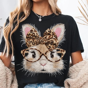 May include: Black t-shirt featuring a bunny face with glasses and a leopard print bow. The bunny has pink ears and is wearing tortoise shell glasses. The design is a whimsical and fun graphic.