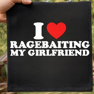 May include: A black square with the phrase "I ❤️ RAGEBAITING MY GIRLFRIEND" in white text. The heart symbol is red. The square is being held by a hand.
