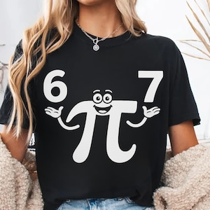 May include: Black t-shirt with a white graphic of the mathematical symbol Pi with arms and a smiling face, between the numbers 6 and 7. The shirt is a crew neck.