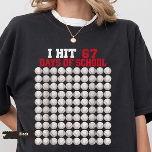 Peut inclure: T-shirt noir avec l'inscription "I HIT 67 DAYS OF SCHOOL" en blanc et rouge. Le motif comprend une grille de balles de baseball. Le t-shirt est de style décontracté.