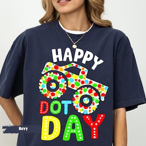 May include: Navy blue t-shirt with the words "HAPPY DOT DAY" in white, red, yellow, and blue. A colorful monster truck graphic with polka dots decorates the shirt. The shirt is a casual style.