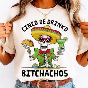 May include: Off-white t-shirt with a Cinco de Mayo theme. The graphic features a skeleton wearing a sombrero and sunglasses, holding a taco and a margarita. Text includes "CINCO DE DRINKO" and "BITCHACHOS".