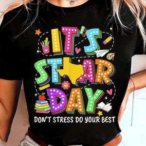 May include: Black t-shirt with a colorful graphic that reads "IT'S STAR DAY" with a Texas-shaped design. The shirt also includes the text "DON'T STRESS DO YOUR BEST" and features school-themed illustrations.