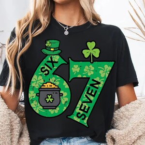May include: Black t-shirt with a green graphic design featuring the numbers "6" and "7" in a shamrock pattern. The number "6" has a leprechaun hat and a pot of gold. The word "SIX" is printed on the number "6" and the word "SEVEN" is printed on the number "7".