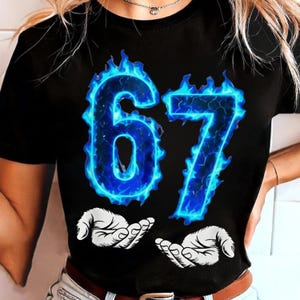 May include: Black t-shirt featuring the number "67" in a vibrant blue flame design. Below the numbers are two white hands with open palms. The shirt is a classic crew neck style.
