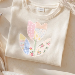 May include: A cream-coloured sweatshirt with a patchwork tulip design. The tulip features pink polka dots, blue dots, yellow, and floral patterns. The stem and leaves are green. The sweatshirt has a crew neck and a small label at the neckline.