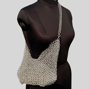 Puede incluir: Bolso de hombro de cota de malla plateada sobre un maniquí negro. El bolso está hecho de pequeños anillos metálicos entrelazados, creando una textura similar a una malla. La correa también es de cota de malla. Tiene forma curva y abierta.