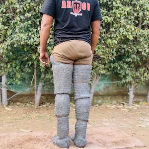 Może przedstawiać: Osoba nosząca zbroję n&oacute;g z kolczugi, brązowy element odzieży i czarny t-shirt ze słowem "ARMOR" w kolorze czerwonym i białym. Kolczuga zakrywa nogi i stopy.