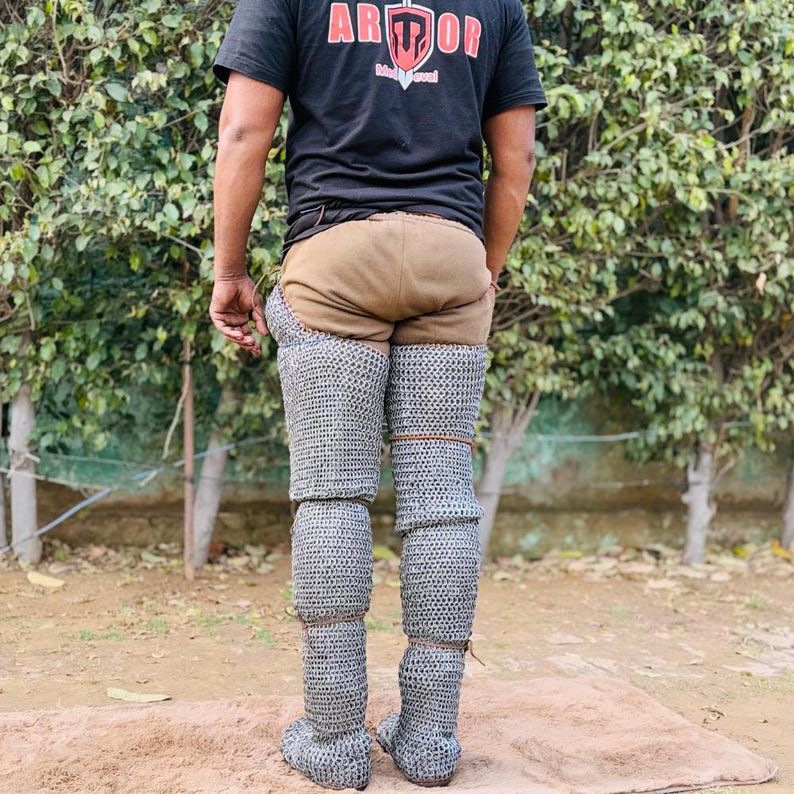 Może przedstawiać: Osoba nosząca zbroję n&oacute;g z kolczugi, brązowy element odzieży i czarny t-shirt ze słowem "ARMOR" w kolorze czerwonym i białym. Kolczuga zakrywa nogi i stopy.