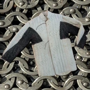 May include: A white quilted gambeson with black chainmail on the shoulders and upper arms. The medieval garment has a stand-up collar and is buttoned down the front. The background is a pattern of metal chain links.