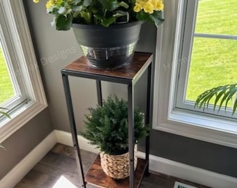 May include: A two-tiered plant stand with a black metal frame and two wooden shelves. The top shelf holds a black pot with yellow flowers. The bottom shelf displays a woven basket with a green plant.