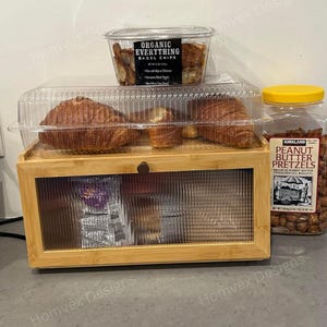 May include: A wooden display case with a clear glass door holds packaged snacks. Above, a clear container of bagel chips and a plastic container of croissants are visible. A jar of peanut butter pretzels with a yellow lid is on the right.