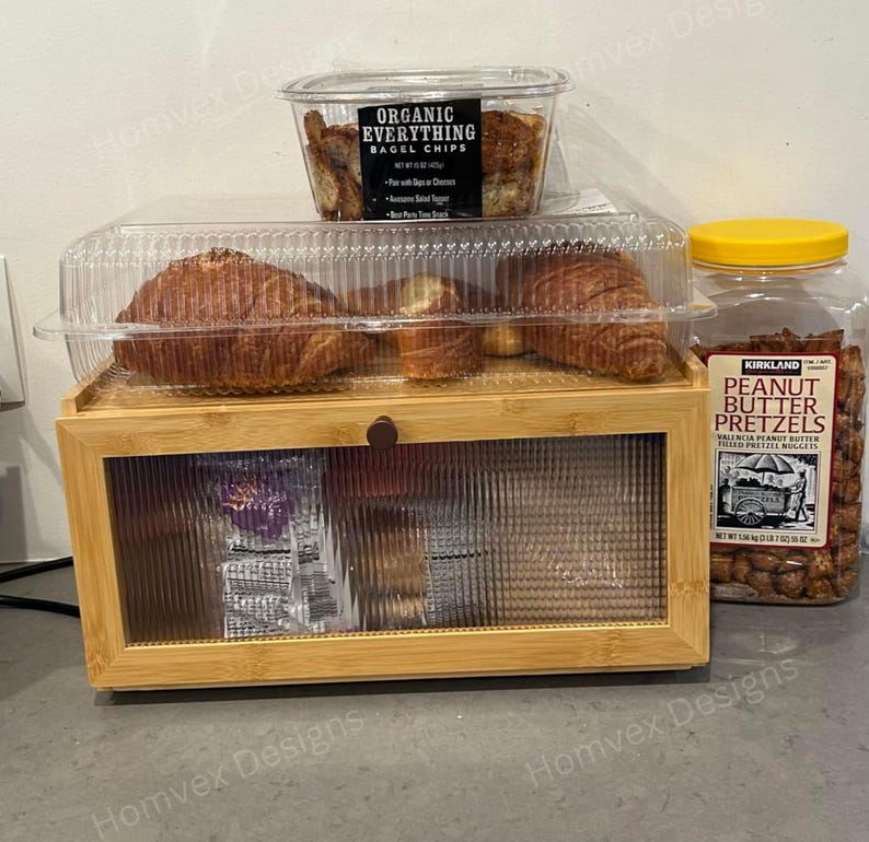 May include: A wooden display case with a clear glass door holds packaged snacks. Above, a clear container of bagel chips and a plastic container of croissants are visible. A jar of peanut butter pretzels with a yellow lid is on the right.