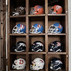 May include: A wooden display case with multiple compartments, each holding a miniature football helmet. The helmets are in various colors, including brown, orange, blue, black, and white, with team logos and numbers. The case has a hinged door.