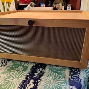 May include: A rectangular bamboo bread box with a clear, ribbed glass front panel and a wooden knob. The top of the box is flat, and the sides are made of bamboo. The box is sitting on a floral patterned tablecloth.
