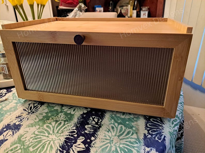 May include: A rectangular bamboo bread box with a clear, ribbed glass front panel and a wooden knob. The top of the box is flat, and the sides are made of bamboo. The box is sitting on a floral patterned tablecloth.