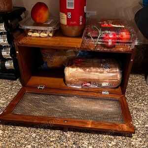 May include: A wooden bread box with a glass front door, filled with bread, tomatoes, and other food items. The box is made of dark wood and has two shelves. A jar of peanut butter and a peach are on top.