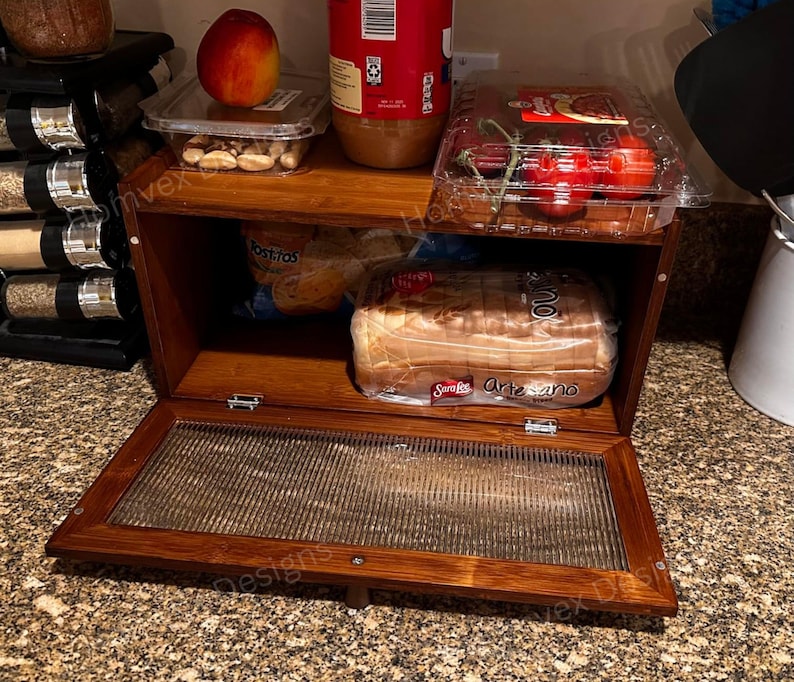 May include: A wooden bread box with a glass front door, filled with bread, tomatoes, and other food items. The box is made of dark wood and has two shelves. A jar of peanut butter and a peach are on top.