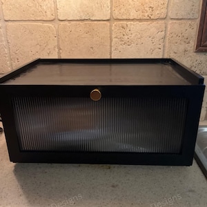 May include: A black cabinet with a ribbed glass door and a gold-colored knob. The cabinet is rectangular and sits on a countertop. The background features a tiled wall.