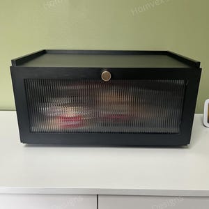 May include: A black storage box with a ribbed glass front and a gold-colored knob. The box has a flat top and is sitting on a white surface. The words "Homvex Designs" are printed on the top and bottom of the image.