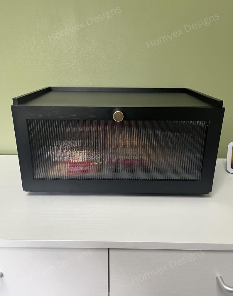 May include: A black storage box with a ribbed glass front and a gold-colored knob. The box has a flat top and is sitting on a white surface. The words "Homvex Designs" are printed on the top and bottom of the image.