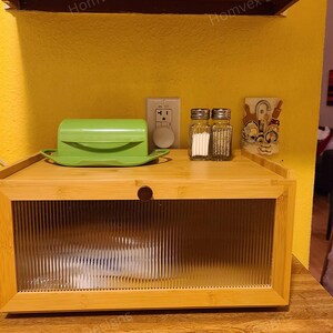 May include: A bamboo bread box with a clear, ribbed glass door. A green butter dish, salt and pepper shakers, and a decorative item sit on top. The background is a yellow wall with an electrical outlet.