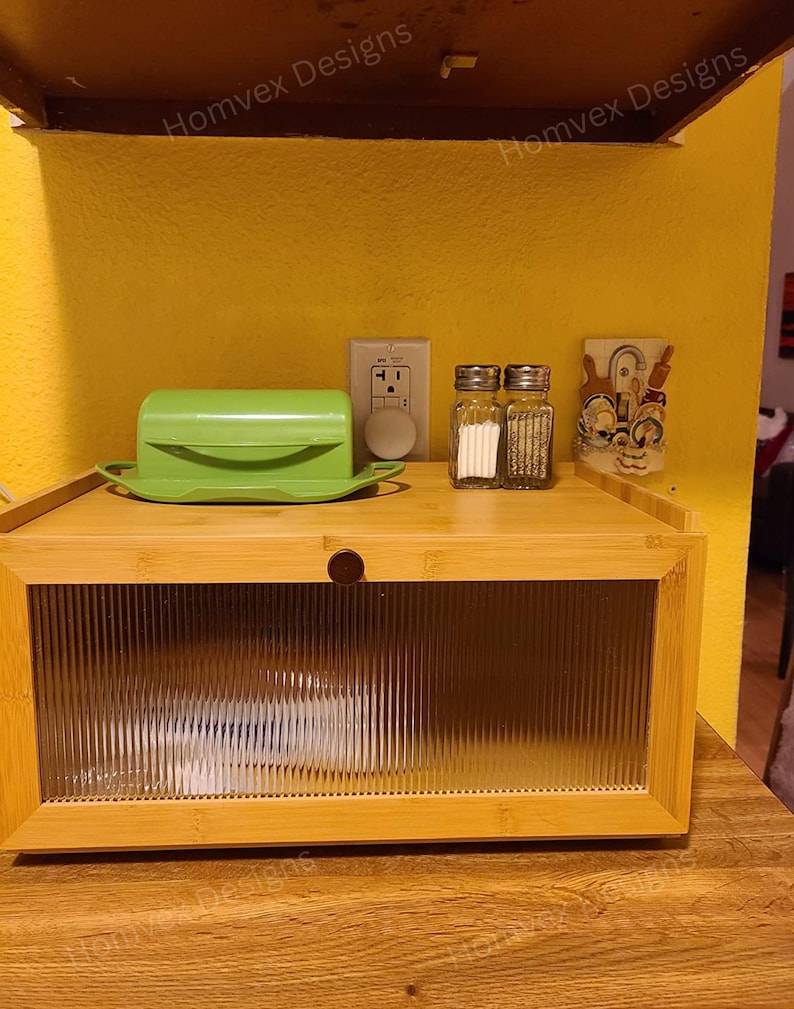 May include: A bamboo bread box with a clear, ribbed glass door. A green butter dish, salt and pepper shakers, and a decorative item sit on top. The background is a yellow wall with an electrical outlet.