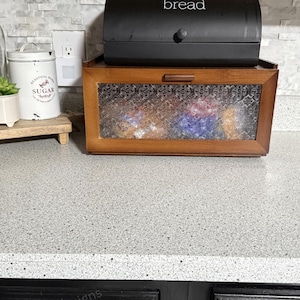 May include: A two-tiered bread box. The top is a black, arched container with the word "bread" in white. The bottom is a wooden box with a glass door. A white container with the word "sugar" is visible.