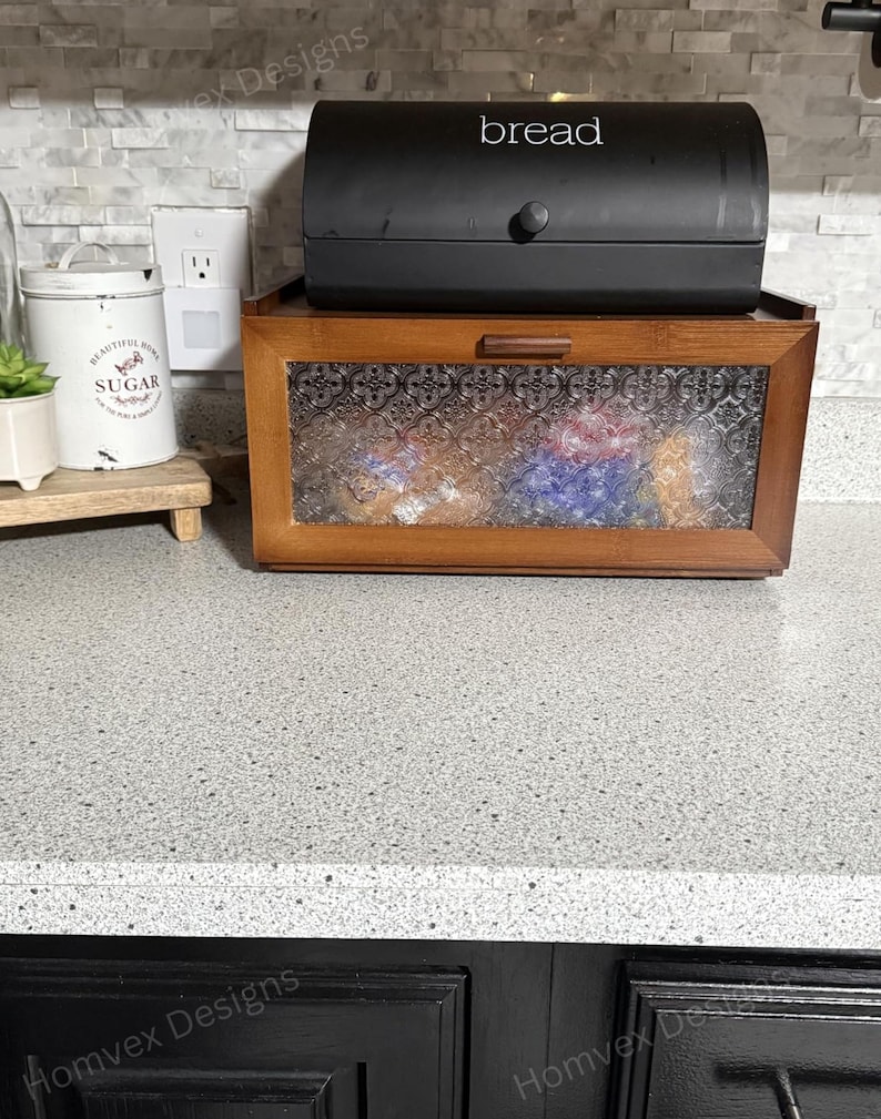 May include: A two-tiered bread box. The top is a black, arched container with the word "bread" in white. The bottom is a wooden box with a glass door. A white container with the word "sugar" is visible.