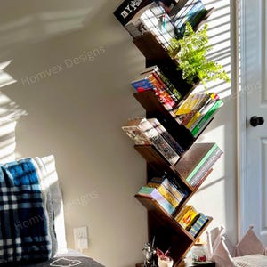 May include: A dark brown, tree-shaped bookshelf filled with books and a small plant. The bookshelf has a unique zig-zag design and sits against a light-colored wall. The top shelf displays the text "MY ROMANCE."