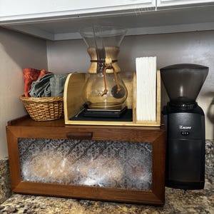May include: A coffee station setup featuring a glass Chemex coffee maker, a coffee grinder, and a wooden breadbox with a patterned glass door. A small basket with towels and a stack of filters are also visible.