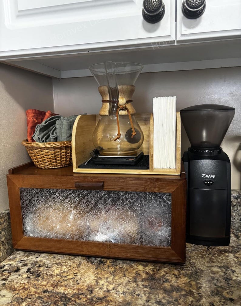 May include: A coffee station setup featuring a glass Chemex coffee maker, a coffee grinder, and a wooden breadbox with a patterned glass door. A small basket with towels and a stack of filters are also visible.