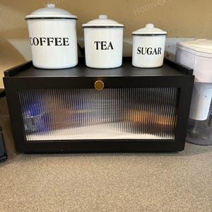 May include: A set of three white enamel canisters labeled "COFFEE", "TEA", and "SUGAR" with black lettering and trim. They sit on a black cabinet with a ribbed glass door and a gold knob. The cabinet is on a countertop.