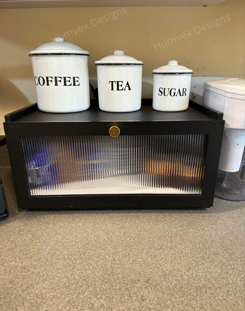 May include: A set of three white enamel canisters labeled "COFFEE", "TEA", and "SUGAR" with black lettering and trim. They sit on a black cabinet with a ribbed glass door and a gold knob. The cabinet is on a countertop.