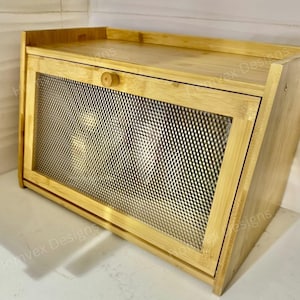 May include: A bamboo bread box with a mesh front door and a wooden knob. The box has a rectangular shape with a slightly angled front. The natural wood color gives it a rustic look.