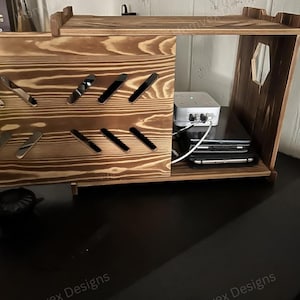May include: A stained wooden charging station with a sliding door and multiple slots for holding devices. The station has a shelf on top and a hexagonal cutout on the side. Inside are electronic devices and a power strip.