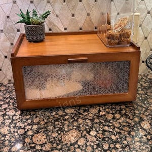 May include: A wooden bread box with a clear, patterned glass front and a small handle. A small succulent plant in a decorative pot sits on top, along with a clear container of baked goods. A black, tiered fruit basket holds bananas and apples.