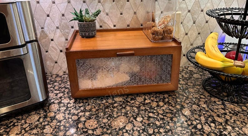 May include: A wooden bread box with a clear, patterned glass front and a small handle. A small succulent plant in a decorative pot sits on top, along with a clear container of baked goods. A black, tiered fruit basket holds bananas and apples.