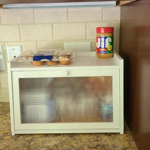 May include: A white bread box with a textured glass door, holding a container of muffins and a jar of Jif peanut butter. The bread box is on a countertop with a neutral-colored backsplash.