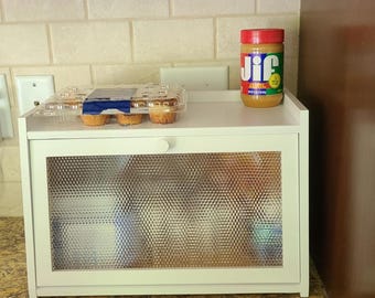 May include: A white bread box with a textured glass door, holding a container of muffins and a jar of Jif peanut butter. The bread box is on a countertop with a neutral-colored backsplash.